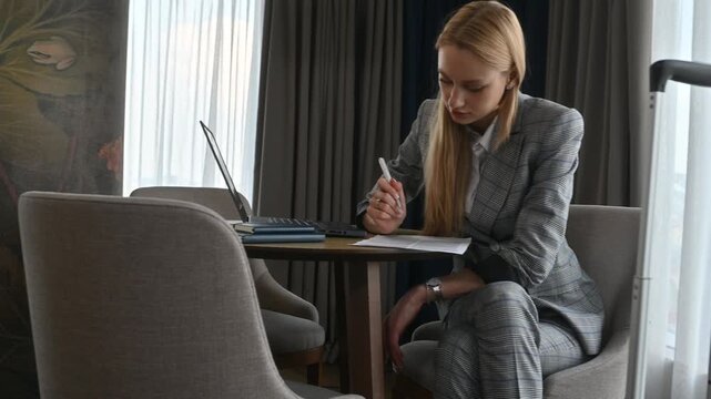 Young businesswoman in a suit sitting at a table in her hotel room, carefully reading and signing a contract, with her laptop and luggage nearby during a business trip