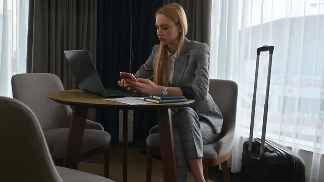 Businesswoman multitasking with her laptop and smartphone while sitting at a table in her hotel room, with her luggage nearby, indicating she is on a business trip