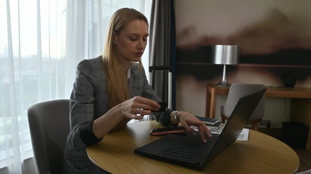 Businesswoman sitting at a table in a hotel room, entering credit card details on her laptop to complete an online purchase, symbolizing modern e-commerce and digital finance