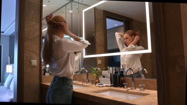 Young woman with long blonde hair styling it into a ponytail while looking at her reflection in a modern bathroom mirror, getting ready for the day ahead in her hotel room