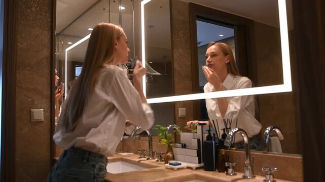 Young woman doing her makeup routine in a luxurious bathroom, applying foundation with a sponge while looking at her reflection in an illuminated mirror before a night out