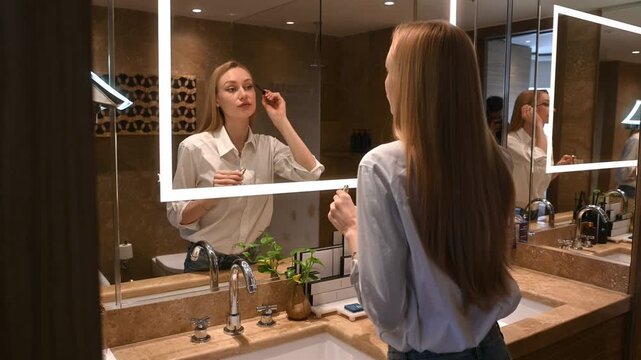 Young woman applying mascara to her eyelashes while looking in an illuminated mirror, getting ready for the day with her daily makeup and beauty routine in a luxury hotel bathroom