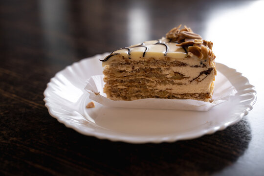 Traditional Esterh&aacute;zy cake served on a white ceramic plate on a wooden cafe table. Close-up food photography featuring layered cake and creamy filling.