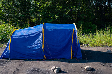 Blue camping tent secured with ropes and rocks on riverside ground near dense forest © Mikhail Mishchenko