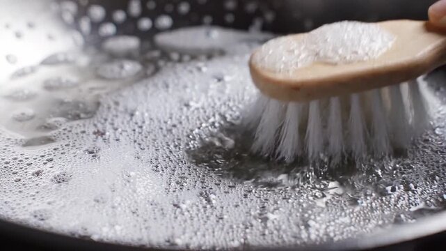Wooden scrub brush cleaning a soapy metal pan, demonstrating household dishwashing and grease removal with foamy water
