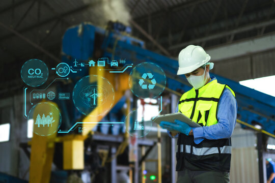 Asian engineer in hardhat using tablet with green energy and carbon neutral HUD graphics inside recycling factory plant.