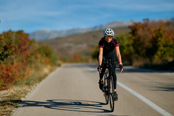 Naklejka premium Female Road Cyclist Training on Open Country Road in Autumn With Helmet and Sunglasses