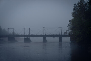Naklejka premium Foggy suspension bridge spanning calm river with muted tones and trees partially visible through dense mist