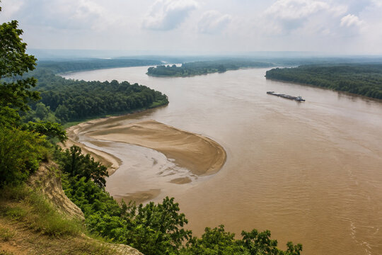 View from high bluff of wide brown river bending through low green forest, sandbar, tugboat and barge downstream, Mississippi River landscape