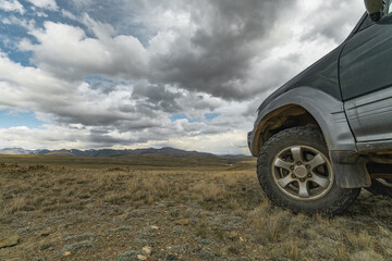 Close up of an off-road suv car wheel with all-terrain tire parked on a grassy hill overlooking a vast mountain valley under a dramatic cloudy sky, overlanding and road trip concept © ANDREY