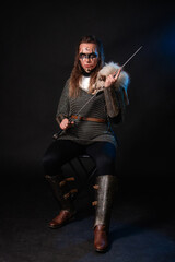 Fototapeta premium Young Viking woman with combat makeup on her face in chainmail with fur and a sword in her hands posing while sitting on a dark background.
