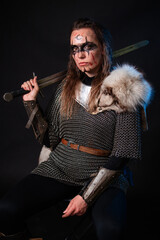 Fototapeta premium Young Viking woman with combat makeup on her face in chainmail with fur and a sword in her hands posing while sitting on a dark background.