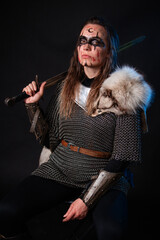 Fototapeta premium Young Viking woman with combat makeup on her face in chainmail with fur and a sword in her hands posing while sitting on a dark background.