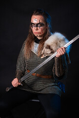 Fototapeta premium Young Viking woman with combat makeup on her face in chainmail with fur and a sword in her hands posing while sitting on a dark background.