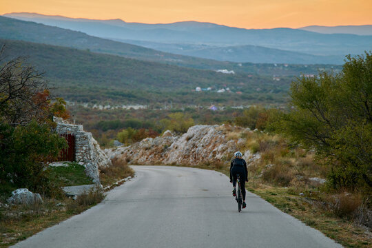 Cyclist Riding On Quiet Country Road At Sunset Through Hilly Countryside Landscape