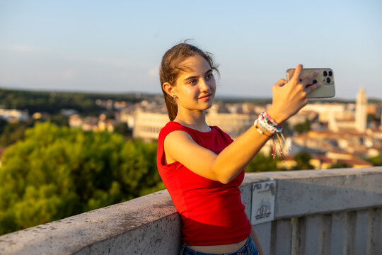Teenage Girl Taking Smartphone Selfie at Pula Viewpoint. Teenage girl in a red top taking a smartphone selfie at a scenic viewpoint overlooking Pula in warm evening sunlight.
