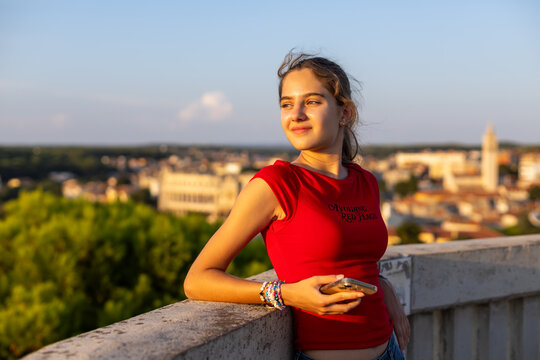Young Girl Holding Smartphone at Sunset City Overlook. Young girl in a red top holding a smartphone and leaning on a concrete railing at sunset with a softly blurred cityscape behind her.