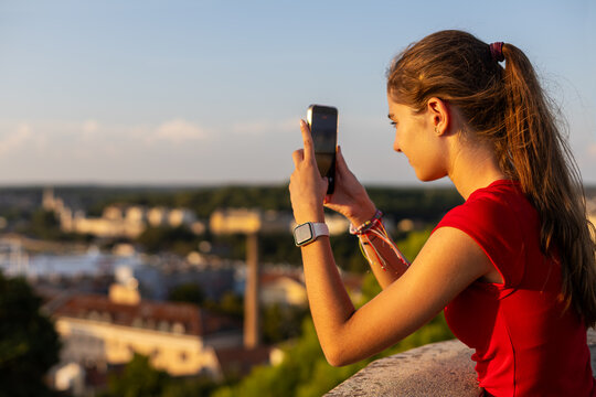 Young Girl Photographing City View with Smartphone at Sunset. Young girl in a red top taking a photo with a smartphone from a terrace at sunset with a blurred city view behind her.