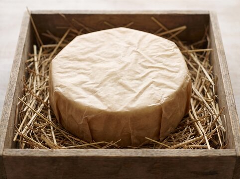 Artisanal cheese wheel wrapped in parchment paper resting on natural straw inside a vintage wooden crate box for gourmet food gift packaging and farm direct delivery