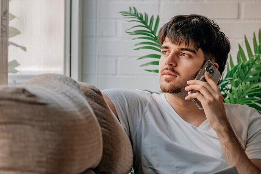 Young man sitting indoors talking on smartphone, looking thoughtfully out window