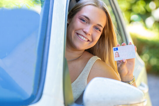Happy young woman leaning out of car window smiling and showing her new driver's license, celebrating passing driving test outdoors