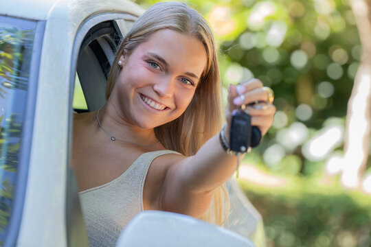 Young smiling woman holding car keys, sitting in her new car with door open, happy driver showing off first vehicle purchase outdoors
