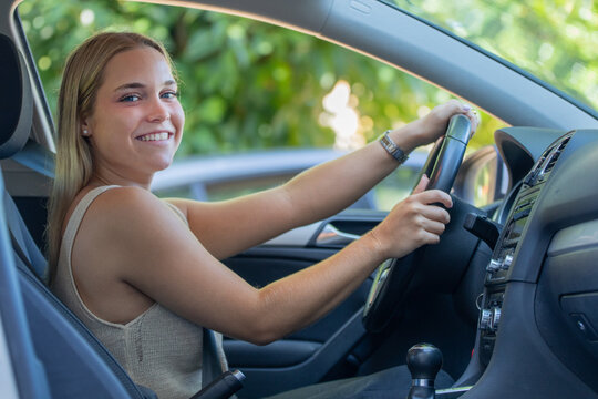 Young woman smiling and looking over her shoulder while driving a car, holding the steering wheel with both hands in a bright, natural outdoor setting