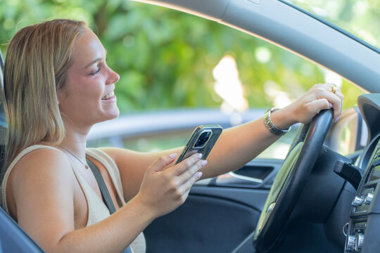 Young smiling woman driving a car and using smartphone, happy female driver texting on mobile phone while holding steering wheel, distracted driving concept
