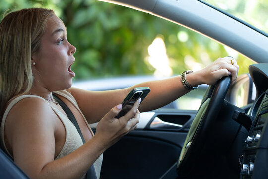 Shocked young woman driving a car while looking at her smartphone, distracted driving concept