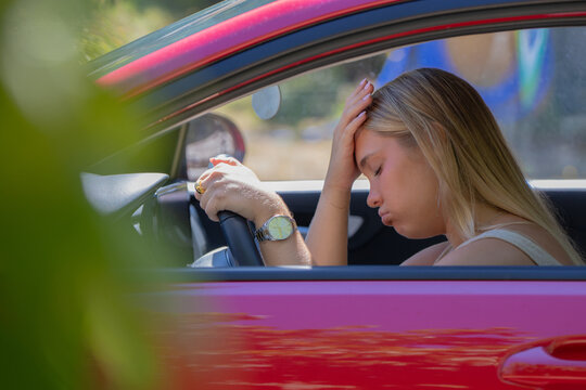 Young woman sitting in car with hand on forehead, looking stressed and exhausted while holding steering wheel