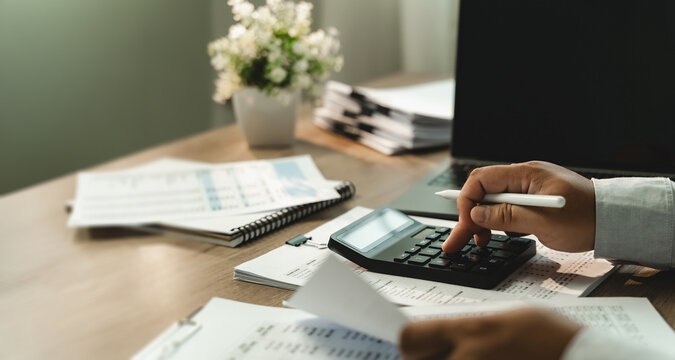 Businessman using calculator over printed financial documents beside laptop for corporate budgeting, accounting review, revenue analysis, expense control, strategic budget planning with copy space.