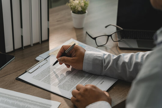 Business person reviewing agreement papers on a clipboard with a visible signature line, eyeglasses and laptop, showing contract signature preparation and approval workflow.