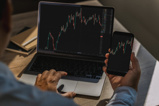 Businessman comparing candlestick stock charts on laptop and smartphone, showing cross-device market analysis, online trading research and investment monitoring.