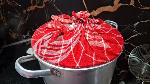 A steamer pot covered with a red and white checkered cloth tied at the top, placed on a stove while steam gently rises from the side.