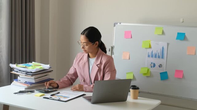 Asian businesswoman suffering from arm, shoulder and neck pain at desk while overworking with laptop and calculator for office syndrome, corporate financial stress and health concept