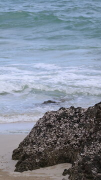 The image features a close up view of a rocky shoreline with dark, rough textured rocks partly embedded in the sand. The rocks are positioned in the foreground, extending towards the water