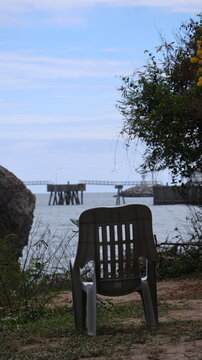 A solitary plastic chair faces a body of water, positioned on a patch of ground with sparse vegetation