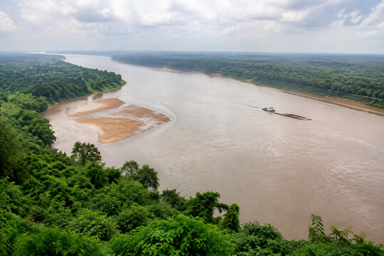 View from high bluff of wide brown river bending through low green forest, sandbar, tugboat and barge downstream, Mississippi River landscape