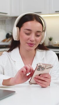 Young woman at home wears wireless headphones types message on smartphone smiles takes break from work. Caucasian girl on sofa sits at table with laptop checks social media chats relaxes calmer mood