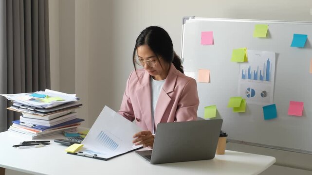 Frustrated Asian businesswoman throwing note paper in anger while working with laptop, calculator and document stack at office desk for corporate stress, mistake and problem concept
