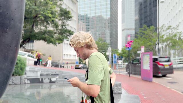 Young Blond Man Using Smartphone on City Street with Skyscrapers Background 