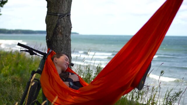 Cinematic footage slowly moving forward to a young woman peacefully napping and cocooning inside a bright orange hammock on a Baltic sea beach after a bicycle ride.