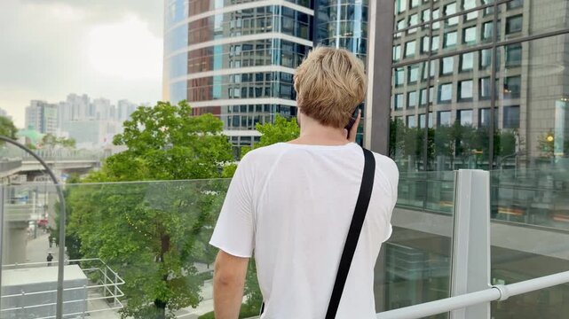 Young Blond Man Standing on Bridge with Smartphone and Skyscraper Skyline in Background &ndash; Summer Day in Modern City