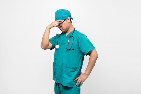 Stressed Asian male doctor in green scrubs touching forehead with headache on white background, Conceptual image representing medical burnout, doctor exhaustion, medical error, and healthcare.