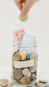 Woman's hand dropping ten Thai Baht coin into a glass jar full of coins. Business and finance concept. Vertical Photography.