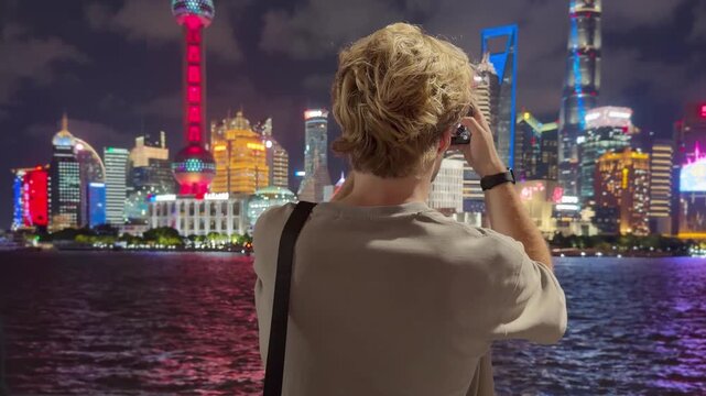Summer Night at The Bund &ndash; Young Blond Man Standing on Waterfront with Smartphone, Reading, Scrolling and Taking Photos Against Skyscraper Skyline in Shanghai