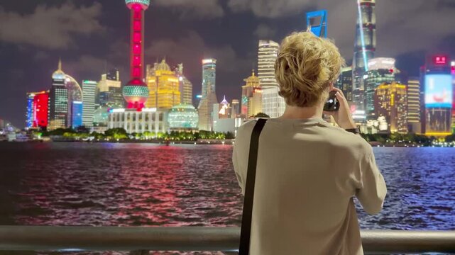 Summer Night at The Bund &ndash; Young Blond Man Standing on Waterfront with Smartphone, Reading, Scrolling and Taking Photos Against Skyscraper Skyline in Shanghai