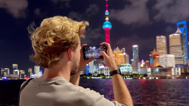 Summer Night at The Bund &ndash; Young Blond Man Standing on Waterfront with Smartphone, Reading, Scrolling and Taking Photos Against Skyscraper Skyline in Shanghai