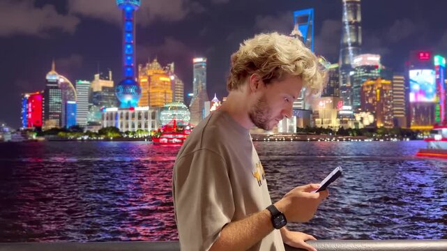 Summer Night at The Bund &ndash; Young Blond Man Standing on Waterfront with Smartphone, Reading, Scrolling and Taking Photos Against Skyscraper Skyline in Shanghai