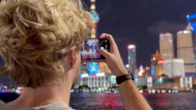 Summer Night at The Bund &ndash; Young Blond Man Standing on Waterfront with Smartphone, Reading, Scrolling and Taking Photos Against Skyscraper Skyline in Shanghai